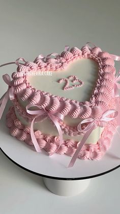 a heart shaped cake with pink icing and ribbons on a white plate in front of a gray background