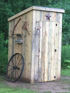 a wooden outhouse sitting in the middle of a field next to a wheel and trees