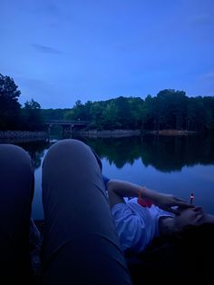 two people laying on the ground next to a body of water with trees in the background