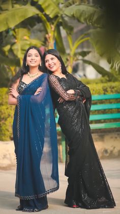 two women standing next to each other in front of some trees and bushes with their arms around one another