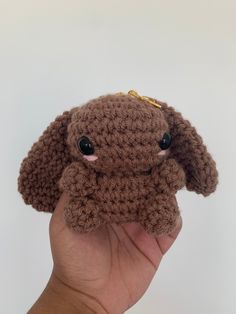 a hand holding a small brown crocheted stuffed animal with eyes on it's head