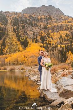 a bride and groom standing on rocks by the water in front of fall colored mountains