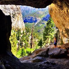 a person standing at the end of a cave looking out on mountains and trees in the distance