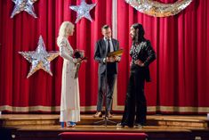 two men and a woman standing on stage in front of red curtains with silver stars