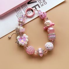 a pink bracelet with charms and beads on top of a table next to a cell phone