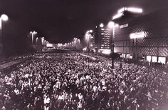 a crowd of people standing on top of a train station platform at night with street lights and buildings in the background