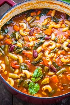 a red pot filled with pasta and vegetables on top of a wooden table next to a green leafy napkin