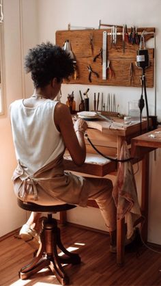 a woman sitting at a wooden desk working on an object with tools hanging on the wall behind her