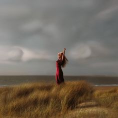 a woman in a long red dress standing on top of a grass covered field next to the ocean