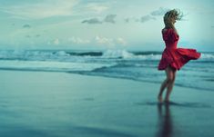 a woman in a red dress is standing on the beach looking at the ocean with her hair blowing in the wind