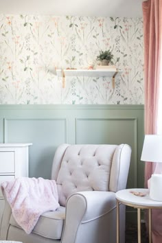 a white chair sitting in front of a window next to a table with a lamp on it