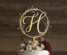 a wedding cake with flowers and a monogrammed letter on top is displayed in front of a wooden wall