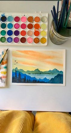 watercolors and other art supplies sit on a desk next to a painting palette