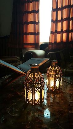 two lit candles sitting on top of a table next to a window with curtains in the background