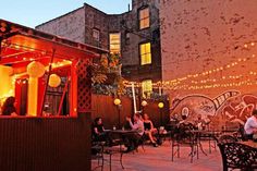 people sitting at outdoor tables in an urban setting with string lights strung across the wall