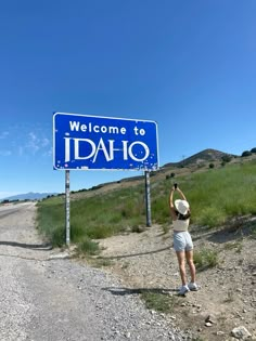 a woman standing in front of a welcome to idaho sign on the side of a road