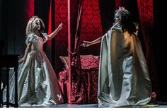 two women dressed in costumes on stage with red curtains behind them and one holding her hand out