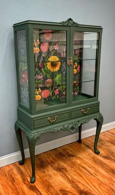 a green china cabinet with flowers painted on the glass doors and bottom drawer is sitting in front of a gray wall