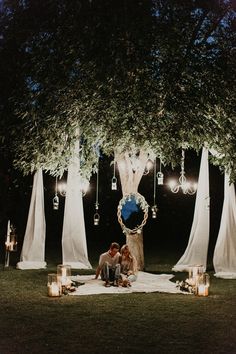 two people sitting under a tree at night with candles in the foreground and lanterns hanging from the trees