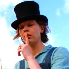 a woman wearing a top hat and holding her finger to her lips while standing in front of a blue sky