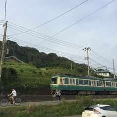 two bicyclists ride past a green train on the tracks in front of a car