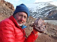 a man in a red jacket is holding up a piece of meat on the side of a mountain