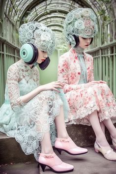 two women wearing headphones and dresses sitting on the ground in front of a fence