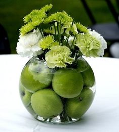 a glass vase filled with green apples and white carnations on top of a table