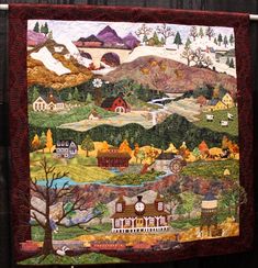 a quilt hanging on the side of a fence with mountains and houses in the background