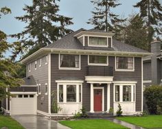 a gray house with white trim and red door in front of some tall trees on the other side