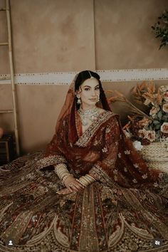 a woman sitting on top of a bed wearing a red and gold wedding dress with flowers in the background