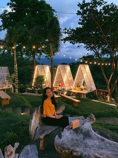 a woman sitting on top of a wooden bench in front of some trees and lights