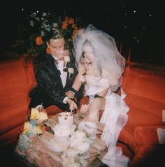 a bride and groom cutting their wedding cake at the same time as they sit on a red couch