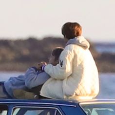 two people sitting on the roof of a car looking at something in front of them