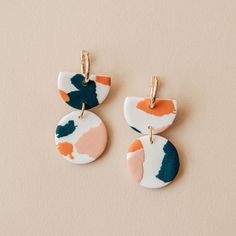 two earrings with different shapes and colors hanging from hooks on a beige surface, the one in the foreground has a gold earring