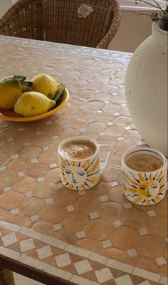 two cups of coffee sitting on top of a table next to lemons and a vase