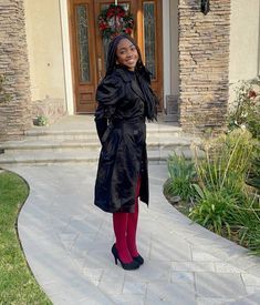 a woman standing in front of a house wearing red tights and black trench coat