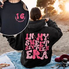 a woman sitting on top of a blanket holding up a black hoodie with pink lettering