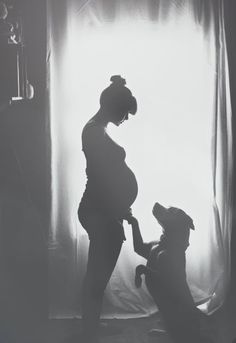 a black and white photo of a pregnant woman with her dog in front of a window