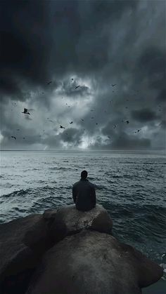 a man sitting on top of a rock next to the ocean under a cloudy sky