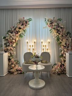 an elegant wedding setup with white chairs and flowers on the back wall, surrounded by candles