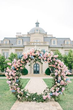 a wedding arch with pink and blue flowers in front of a large mansion on a cloudy day