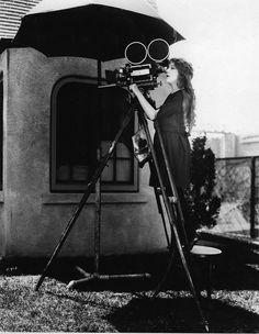 a woman standing in front of a house holding an umbrella and looking through a camera