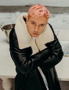 a man with pink hair wearing a black leather jacket and white fur collar standing in front of a counter