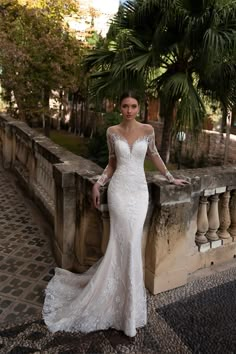 a woman in a wedding dress leaning on a wall with palm trees behind her and looking at the camera