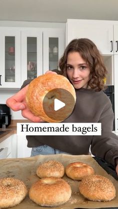 a woman holding a bagel in front of some doughnuts on a baking sheet