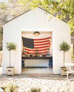 an american flag hanging on the side of a white shed with potted plants and candles