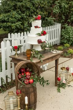 a wedding cake with apples and greenery sits on top of a barrel in front of a white picket fence