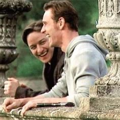 two men sitting next to each other on top of a stone wall with an ornate fountain in the background