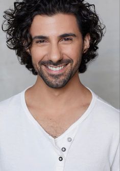 a man with curly hair and a beard smiles at the camera while wearing a white shirt